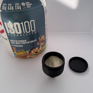 May include: A white and blue protein powder container with the text "ISO100 HYDROLYZED" and "Fruity Pebbles" flavor. A small black container with a scoop of light-colored powder sits next to it.