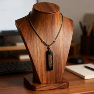 May include: A wooden jewellery display bust holds a necklace. The necklace has a dark green cord, gold accents, and a black rectangular pendant with gold text. A red bead is between the pendant and cord.