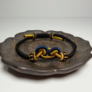 May include: A black and gold bracelet with a knot design. The bracelet features a central knot in navy blue and gold, with gold accents and black cord. The bracelet is displayed on a dark brown, textured plate.