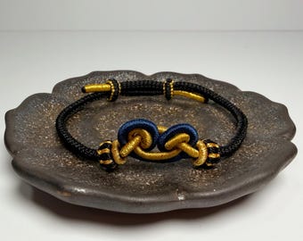 May include: A black and gold bracelet with a knot design. The bracelet features a central knot in navy blue and gold, with gold accents and black cord. The bracelet is displayed on a dark brown, textured plate.