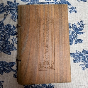 Puede incluir: Un libro de madera con un diseño tallado y texto en la portada. El libro está encuadernado con un cordón marrón oscuro. La portada es de color marrón cálido con vetas de madera visibles. El texto está en formato vertical.