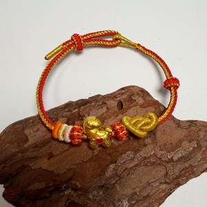 May include: A gold and red braided bracelet with a gold horse charm, heart-shaped knot, and decorative knots. The bracelet is displayed on a piece of wood.