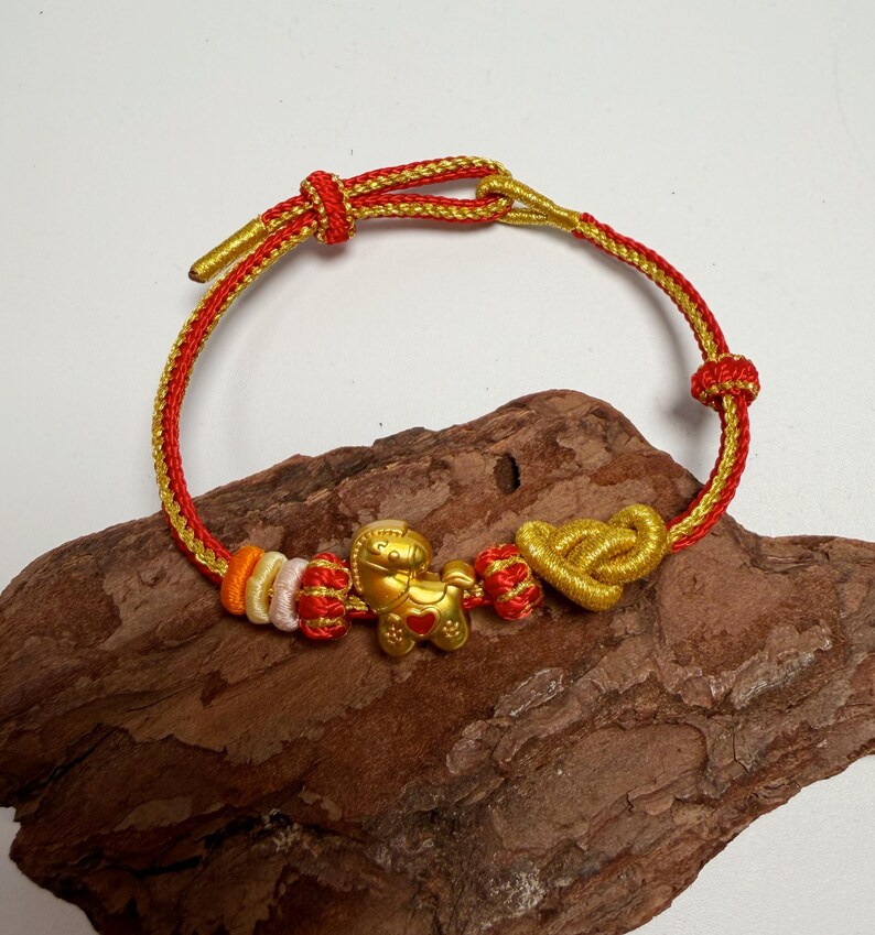 May include: A gold and red braided bracelet with a gold horse charm, heart-shaped knot, and decorative knots. The bracelet is displayed on a piece of wood.