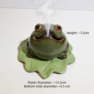 Puede incluir: Un quemador de incienso de cerámica con forma de rana verde, sobre una base en forma de hoja verde. Los ojos de la rana son marrones y sale humo de su boca. Altura aproximada 7,2 cm, diámetro de la placa 13,5 cm.