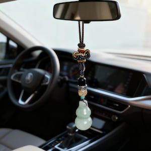 Puede incluir: Interior de coche con un adorno colgante decorativo. El adorno presenta un colgante de jade en forma de calabaza, una cuenta negra y un nudo decorativo con detalles en marrón, dorado y azul. El adorno cuelga del espejo retrovisor.