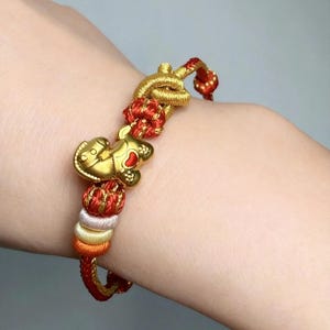 May include: Gold charm bracelet with a red and gold woven cord. The bracelet features a gold charm shaped like a small animal with a red heart. The cord is accented with red, gold, orange, and white beads.