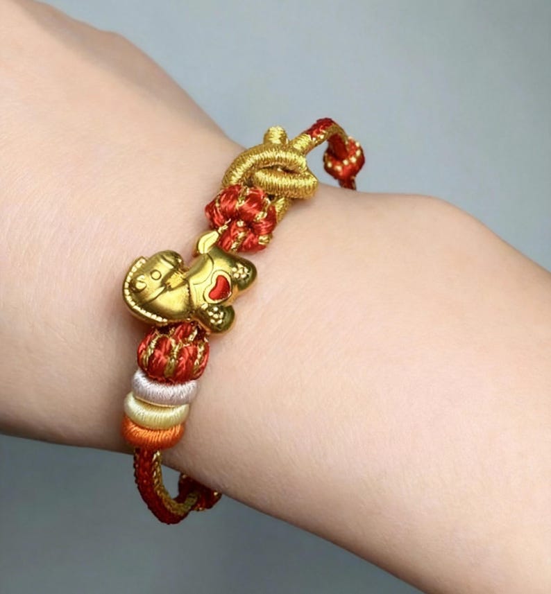 May include: Gold charm bracelet with a red and gold woven cord. The bracelet features a gold charm shaped like a small animal with a red heart. The cord is accented with red, gold, orange, and white beads.