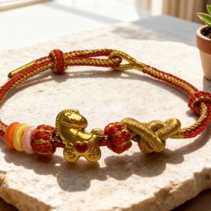 May include: A gold and red braided bracelet with a gold horse charm. The bracelet features a series of colourful beads and intricate knots. The bracelet is displayed on a textured surface.