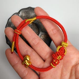May include: Red and gold bracelet featuring a small gold turtle charm. The bracelet is made of woven red cord with gold accents at the clasp and decorative elements. The turtle charm is a focal point of the bracelet.