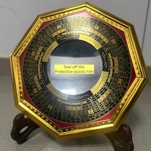 May include: An octagonal, gold-framed compass with a red border and intricate black and gold detailing. The center features a reflective surface covered by a protective film with the text "Tear off the Protective plastic film". It rests on a brown stand.