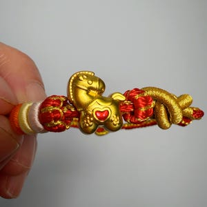 May include: A gold-coloured horse charm bracelet with red and gold braided accents. The horse charm has a red heart-shaped detail. The bracelet is made of braided cord in shades of red, gold, orange, and white.