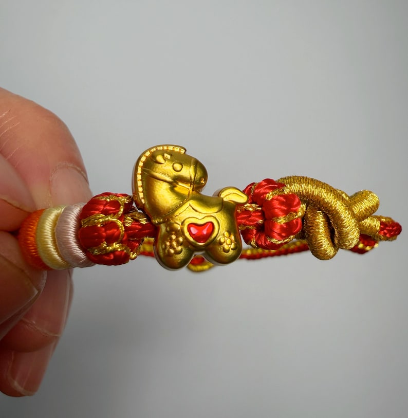 May include: A gold-coloured horse charm bracelet with red and gold braided accents. The horse charm has a red heart-shaped detail. The bracelet is made of braided cord in shades of red, gold, orange, and white.