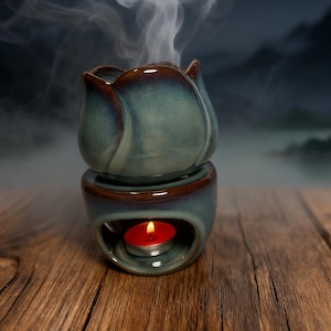 Könnte beinhalten: Ein türkisfarbener Keramik-Aroma-Diffusor in Tulpenform mit braunem Rand und Sockel. Eine brennende rote Kerze befindet sich im Inneren des Sockels, und weißer Dampf steigt von oben auf. Der Diffusor steht auf einer Holzoberfläche.