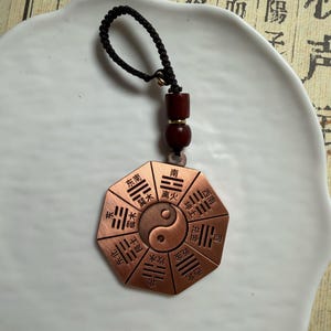 May include: A copper-toned, octagonal pendant with a Yin and Yang symbol at its core. The pendant features Chinese characters and is suspended by a black cord with wooden beads. It is likely a decorative item or charm.
