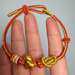 May include: A red and gold bracelet with a gold horse charm and a gold heart charm. The bracelet is made of woven cord and features a sliding knot closure. The bracelet is displayed on a hand.