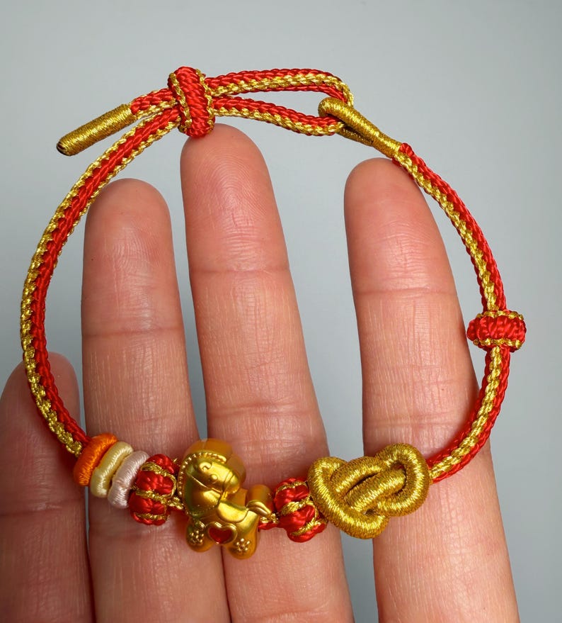 May include: A red and gold bracelet with a gold horse charm and a gold heart charm. The bracelet is made of woven cord and features a sliding knot closure. The bracelet is displayed on a hand.