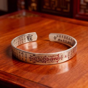 Peut inclure: Bracelet manchette argenté avec caractères chinois gravés et accents rouges. Le bracelet ouvert est présenté sur une surface en bois, mettant en valeur les détails du design. Un bijou unique.
