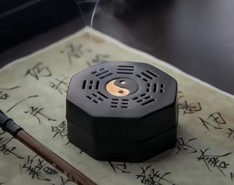 May include: A black, octagonal incense burner with a gold-colored Yin and Yang symbol on top. The burner has a series of small rectangular vents around the top. Smoke rises from the burner, suggesting it is in use. The burner sits on a paper with calligraphy.