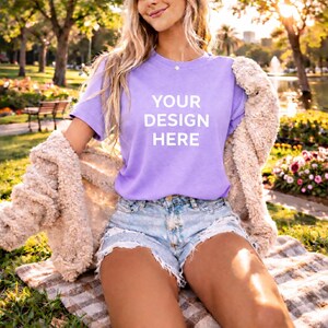 May include: A lilac t-shirt with the text "YOUR DESIGN HERE" in white. The model is wearing denim shorts and a beige jacket. The image is taken outdoors on a sunny day.
