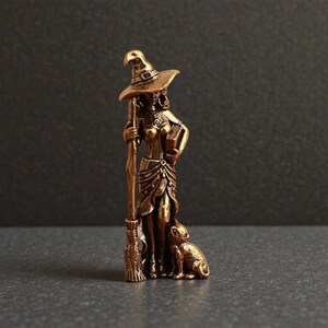 May include: A bronze-colored figurine of a witch wearing a pointed hat, holding a broom and a book. A small dog sits at her feet. The witch is dressed in a detailed outfit. The figurine has a vintage or antique appearance.