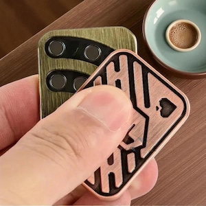 Può includere: Una mano tiene in mano due oggetti metallici. Uno è una custodia per telefono rettangolare verde oliva con tre lenti circolari per fotocamera. L'altro è un giocattolo fidget quadrato color rame con un disegno di carte da gioco. Un piccolo piatto blu-verde con una sostanza simile al caffè è sullo sfondo.
