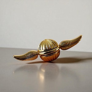 以下が含まれることがあります： 金色のスニッチのレプリカ。2つの繊細な翼が付いた球形のオブジェクトです。ボールはリブ状のテクスチャで、翼は羽の模様で詳細に描かれています。オブジェクトは反射する表面に表示されています。