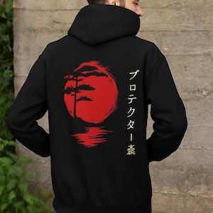 May include: Black hoodie featuring a red graphic of a tree silhouette in front of a red sun, with Japanese text on the right side. The design is reflected in a body of water. The hoodie is worn by a person.