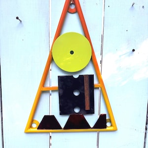 May include: Abstract geometric wall art. A yellow and orange triangle frame holds a chartreuse circle, a black square, and maroon shapes. The artwork is mounted on a light blue wooden surface.
