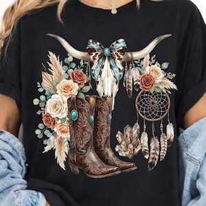 Puede incluir: Camiseta negra con un diseño de temática western. El diseño incluye una calavera de toro con detalles turquesa, elementos florales y botas de vaquero marrones. Un atrapasueños y cristales completan el diseño.