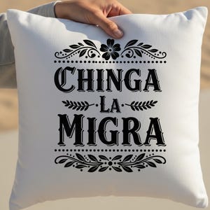 May include: White square pillow with black text and decorative floral elements. The text reads "CHINGA LA MIGRA" in a bold, stylized font, with floral and dotted border designs above and below the text.