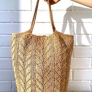 May include: A natural-toned woven tote bag featuring a leaf design. The bag has two long handles and is held up by a hand. The bag is made from a natural fibre and is suitable for carrying items. The bag is a neutral colour.