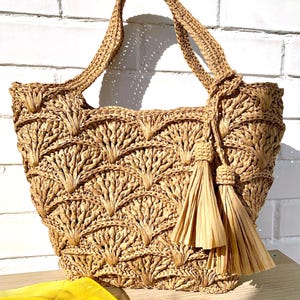 Op de afbeelding: Een geweven raffia-draagtas met een natuurlijke, lichtbruine kleur. De tas heeft een gedetailleerd, boomachtig patroon en twee decoratieve kwastjes. De handvatten zijn ook geweven. Een gele sjaal is zichtbaar op de voorgrond.