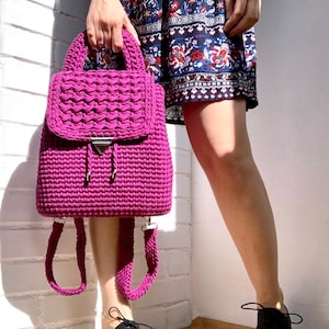 Könnte beinhalten: Ein magentafarbener Häkelrucksack mit Klappenverschluss und verstellbaren Trägern. Die Tasche hat ein strukturiertes Stichmuster und einen silberfarbenen Verschluss. Der Rucksack wird vor einer weißen Backsteinmauer gehalten.