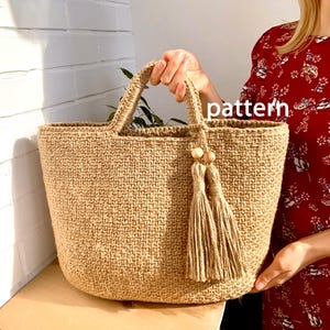 May include: A woven jute tote bag with sturdy handles and a decorative tassel. The bag is a natural tan color and has a rectangular shape. The word "pattern" is visible on the bag.