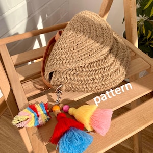 May include: A woven beige handbag with a leather strap, resting on a wooden chair. The bag is accompanied by a keychain with colorful tassels in red, blue, yellow, and pink. The word "pattern" is visible.