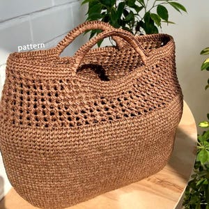 Puede incluir: Un bolso tote marrón grande, con tejido abierto y dos asas resistentes. El bolso está hecho de material tejido, con un aspecto texturizado. La palabra "pattern" es visible en la parte superior izquierda del bolso. Está sobre una superficie de madera.