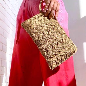 May include: A woven straw clutch bag with a rectangular shape and a braided handle. The bag has a detailed, textured pattern in shades of tan and beige. The background includes bright pink trousers and a white top.
