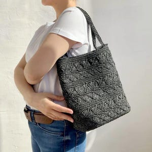 Puede incluir: Un bolso tote gris oscuro con un diseño tejido y texturizado. El bolso tiene forma rectangular y una correa para el hombro. El patrón del bolso presenta un diseño geométrico repetitivo. El bolso lo lleva una persona que lleva una camiseta blanca y vaqueros azules.