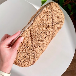May include: A woven straw clutch with a diamond pattern and a silver zipper. The clutch is a light tan color and appears to be handmade. The bag is on a white surface.