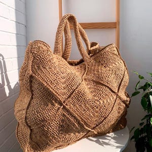 Pode incluir: Uma grande sacola tote tecida em cor bege natural. A sacola apresenta um design de padrão de diamante e alças resistentes. Está em cima de uma mesa branca, com uma escada de madeira ao fundo.