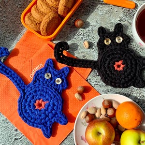 May include: Two handmade crochet cat coasters, one blue and one black, with button eyes and a starburst design. The blue cat has a long tail. The coasters are on an orange napkin with the word "pattern".