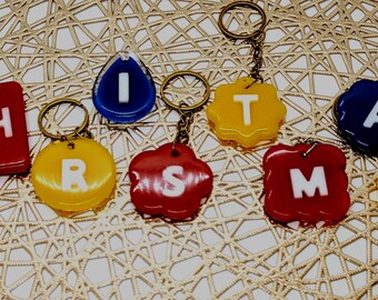 May include: A collection of colorful keychains spelling out "CHRISTMAS". Each keychain features a different letter in white against a background of blue, red, and yellow. The keychains are attached to gold-colored key rings.