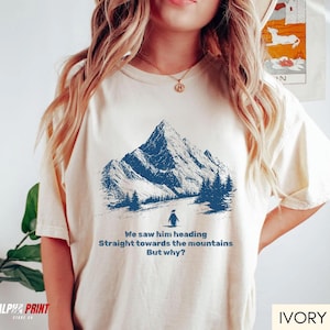 May include: Ivory-colored t-shirt featuring a blue mountain graphic with a penguin and text. The text reads "We saw him heading Straight towards the mountains But why?" The shirt has a relaxed fit and is made of soft material.