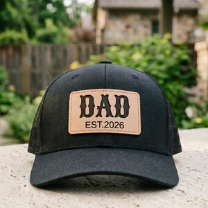 May include: Black trucker hat with a mesh back and a curved brim. The hat features a tan leather patch with the word "DAD" in a bold, decorative font, and the text "EST.2026" below it. The hat is a classic accessory.