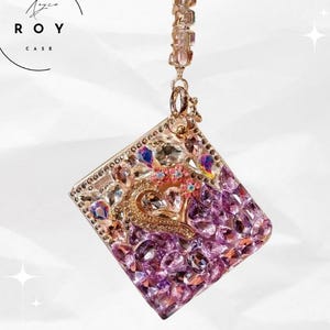 May include: A square phone case with a purple and clear crystal design. The case features a gold-coloured heart-shaped embellishment with a crown and small pink flowers. The case is suspended by a gold-coloured chain.