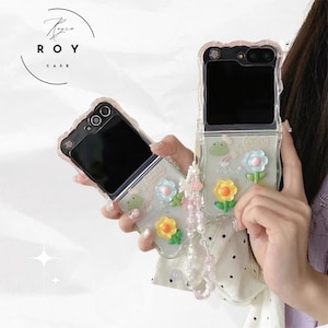 Pode incluir: Capas de telefone transparentes com desenhos florais e de rãs. As capas têm uma borda rosa e são decoradas com flores 3D amarelas e azuis. As capas estão abertas, revelando os ecrãs dos telefones. Uma alça de contas rosa pende de uma capa.