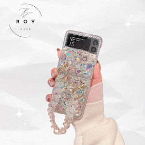 Puede incluir: Una funda transparente para teléfono incrustada con pedrería iridiscente. La funda tiene aros dorados y una correa de cuentas a juego. La pantalla del teléfono muestra la hora como 4:15. La marca "ROY CASE" está impresa en un logotipo circular.