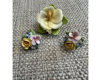 Puede incluir: Tres esculturas florales de porcelana. La más grande es un hibisco color crema con centro amarillo y hojas verdes. Dos arreglos más pequeños presentan rosas amarillas, flores rosas y azules y hojas verdes. Las esculturas están sobre una superficie texturizada de tono neutro.