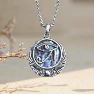 Könnte beinhalten: Eine silberfarbene Anhängerkette mit einem kreisförmigen Design, das ein Auge des Horus-Symbol über einer schillernden Abalone-Muschel-Einlage zeigt. Der Anhänger hängt an einer Boxkette und hat gravierte Flügelmotive an der Basis.