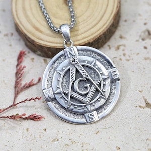 Pode incluir: Colar com pingente prateado com um símbolo maçónico. O pingente apresenta um design de bússola e esquadro com as letras "G", "N", "E", "S" e "W". O colar está em uma corrente prateada e repousa sobre uma superfície de madeira.